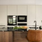 Modern luxury lodge kitchen with black stone island, integrated appliances, pale cabinetry and tan leather breakfast bar chairs.