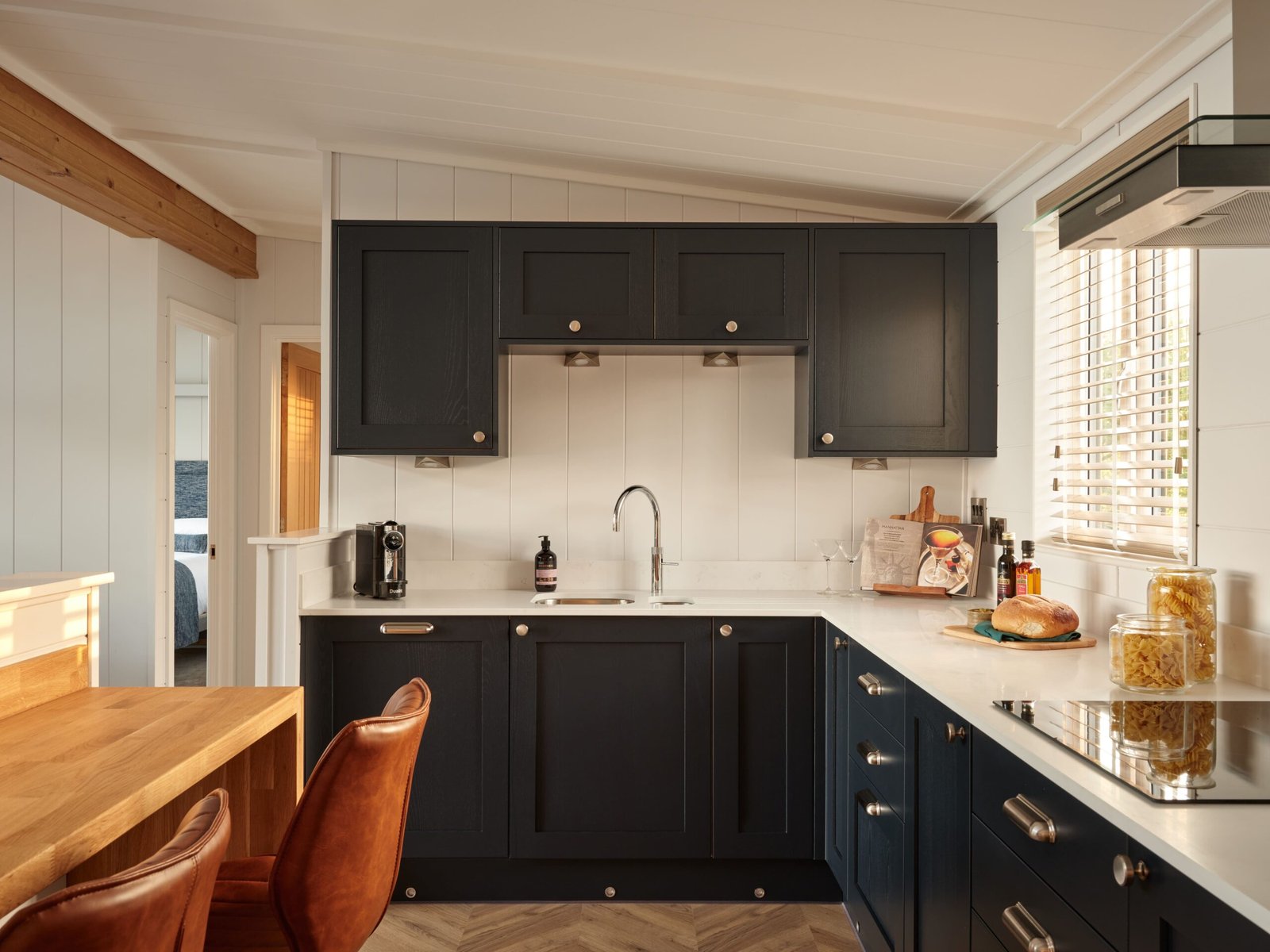 Compact luxury lodge kitchen with dark shaker-style cabinets, white worktops, wooden breakfast table and leather chairs.