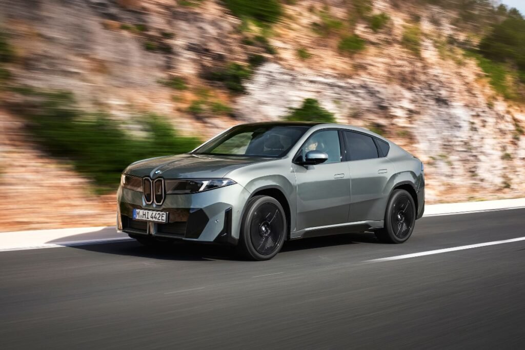 BMW iX4 – Driving Exterior BMW iX4 electric SUV driving on the road, viewed from a front three-quarter angle.