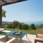 Shaded villa terrace with contemporary outdoor armchairs and a coffee table overlooking the Sicilian coastline.