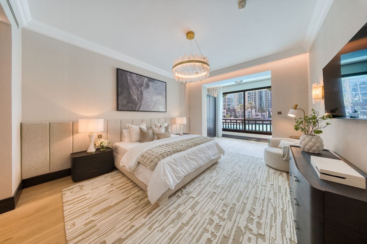 Spacious bedroom with an upholstered bed, chandelier and balcony doors overlooking the Downtown Dubai waterfront.